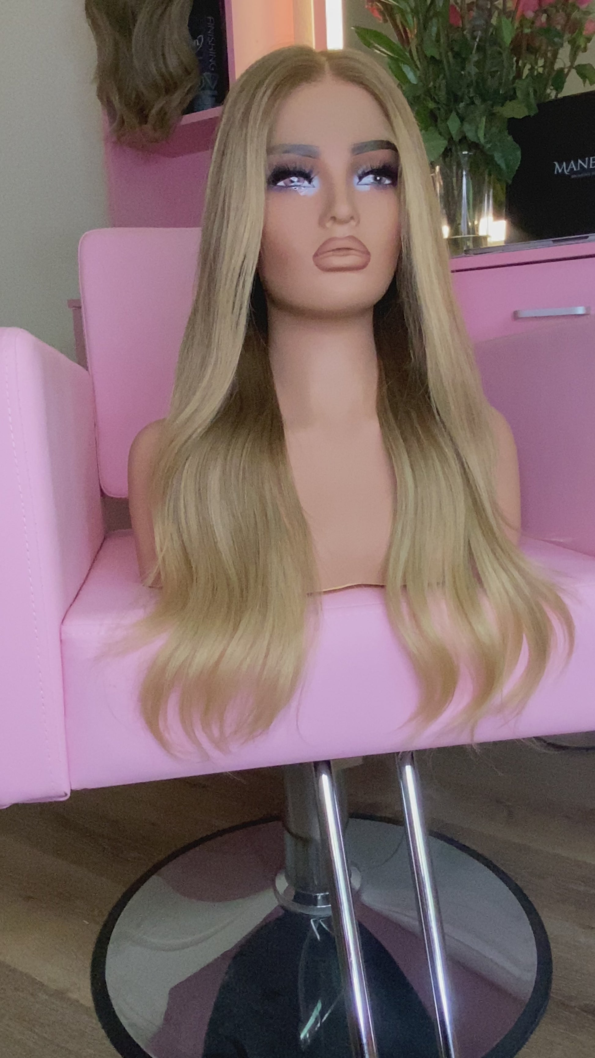 Blonde wig on a mannequin head with 'Mane Wishes' branding, seated on a pink chair.